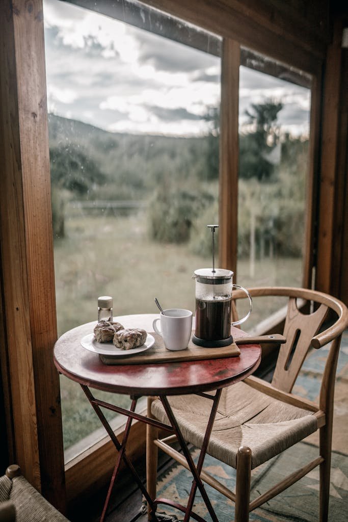 A rustic breakfast scene with coffee and pastries by a large window overlooking nature.