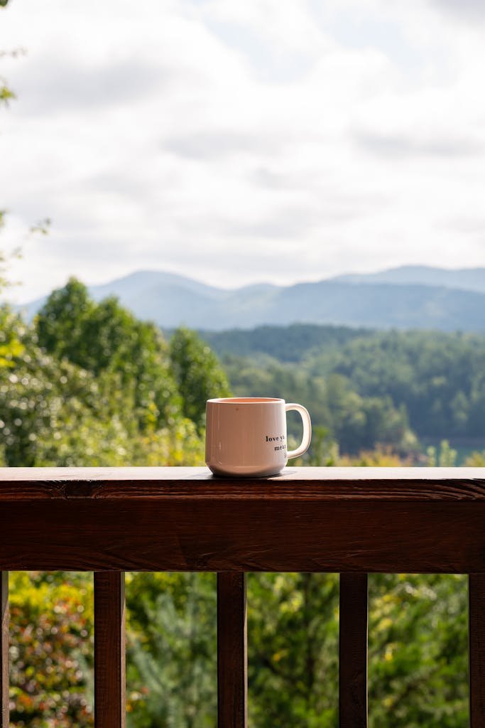 A white coffee mug sits on a wooden railing, with a scenic mountain view in the background.
